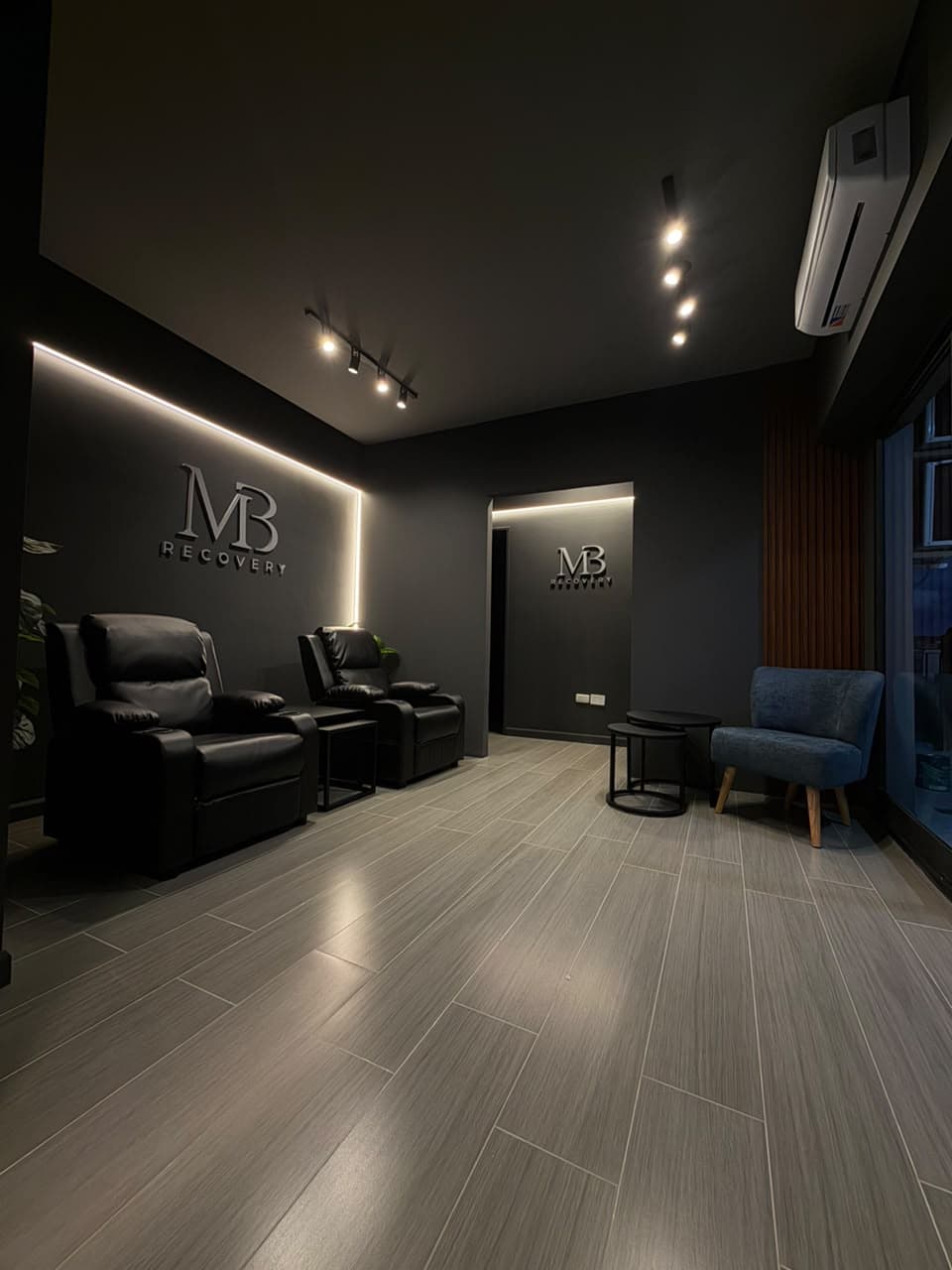 El hall de MB Recovery: el logo iluminado por detrás sobre la pared oscura, dos sillones negros de recuperación a la izquierda, y a la derecha un sillón y mesas bajas junto al ventanal.