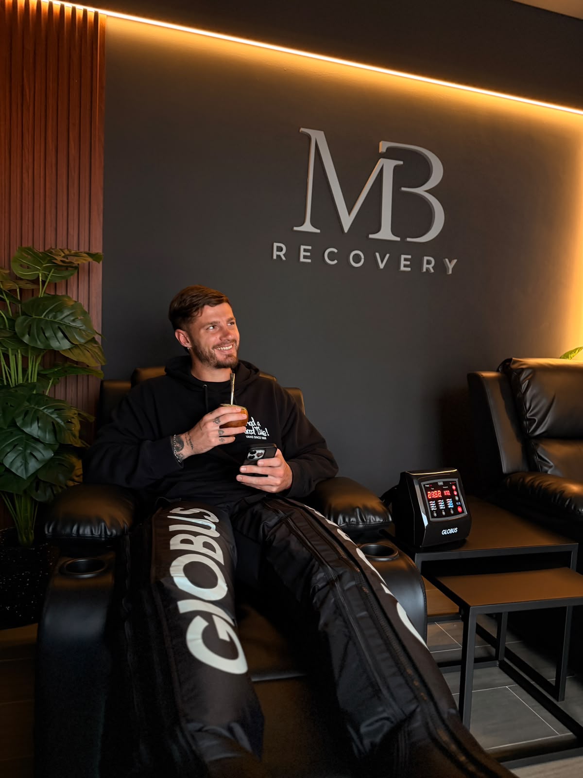 Una persona sentada en uno de los sillones de MB Recovery con las botas de presoterapia puestas hasta la cadera, el equipo de control a su lado y el logo de la marca en la pared.