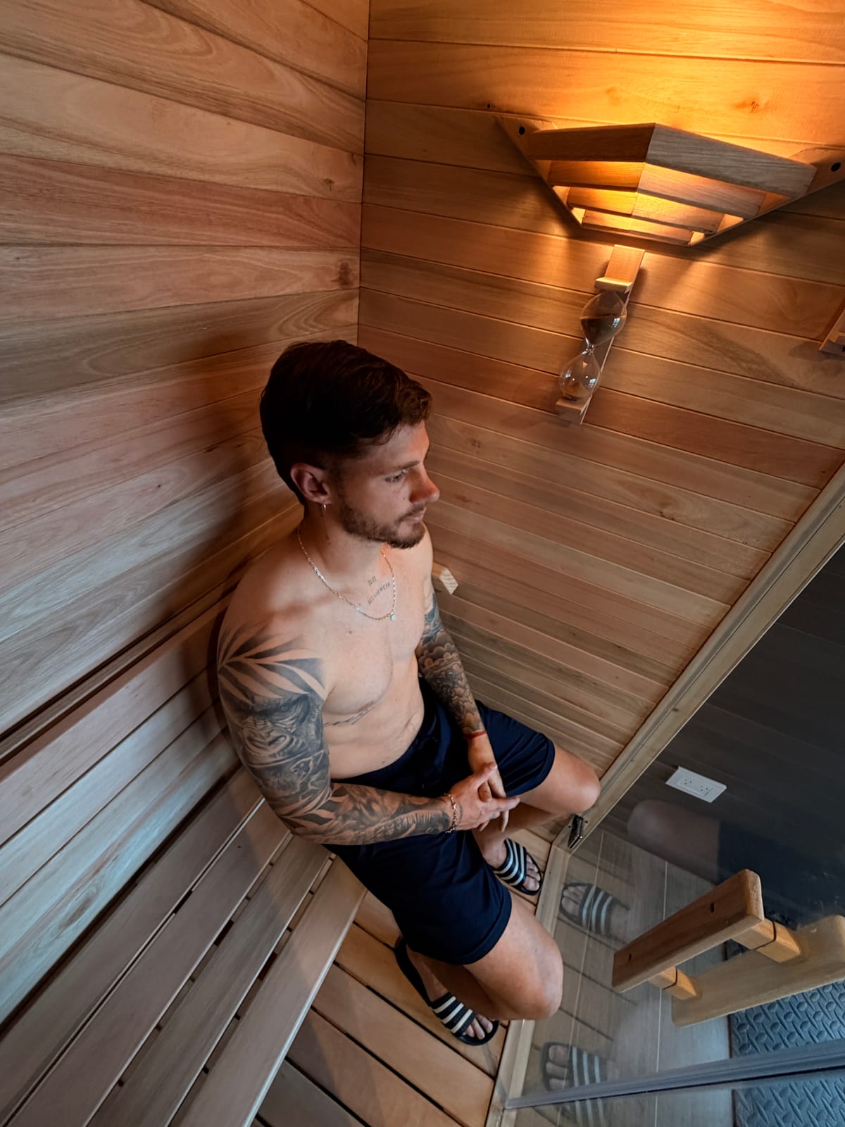 El sauna de MB Recovery visto desde arriba: una persona sentada en el banco de madera, con la luz cálida del techo y un reloj de arena en la pared.